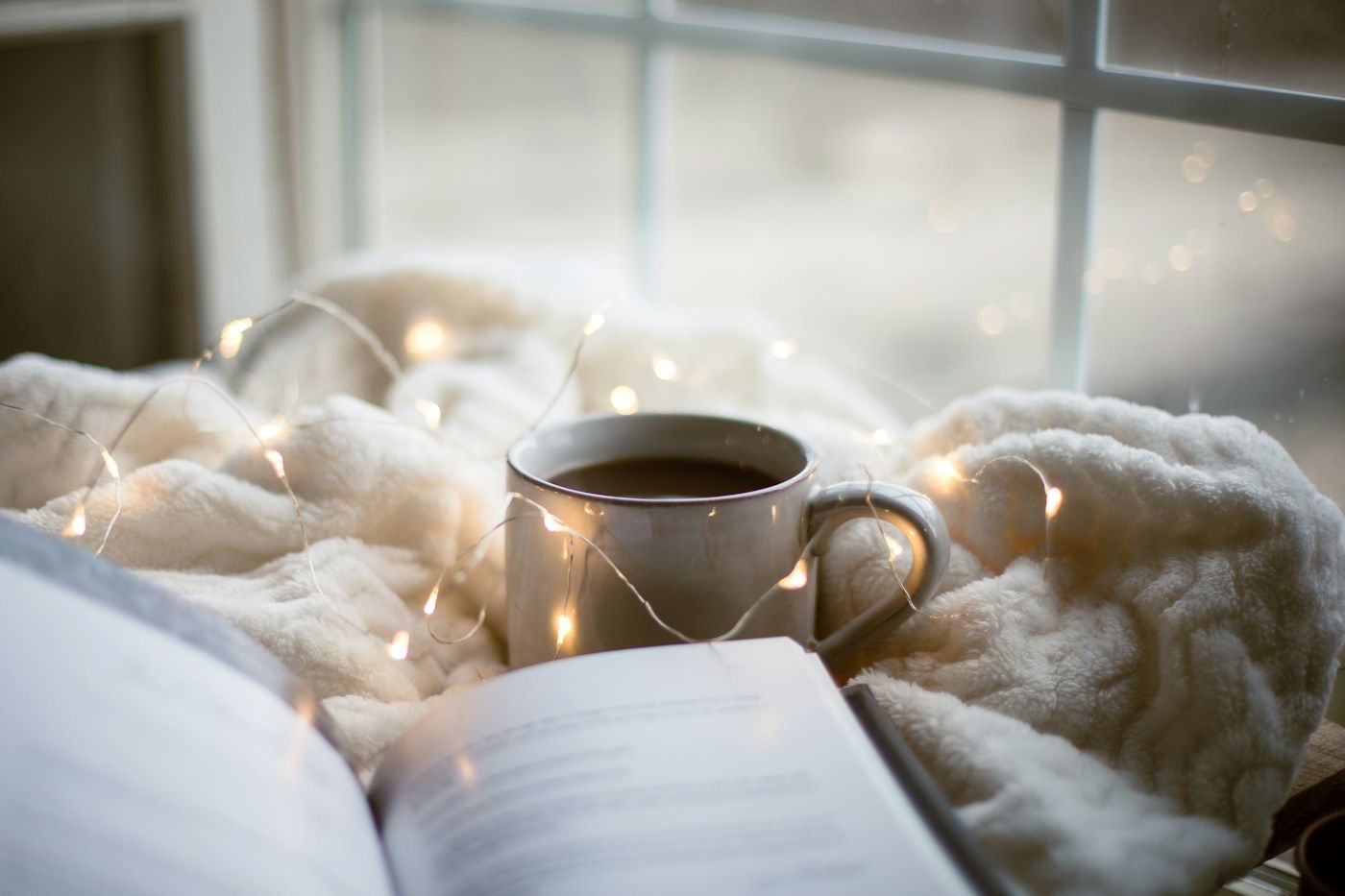 Tasse fumante et livre ouvert sur un plaid en laine près d'une fenêtre d'hiver